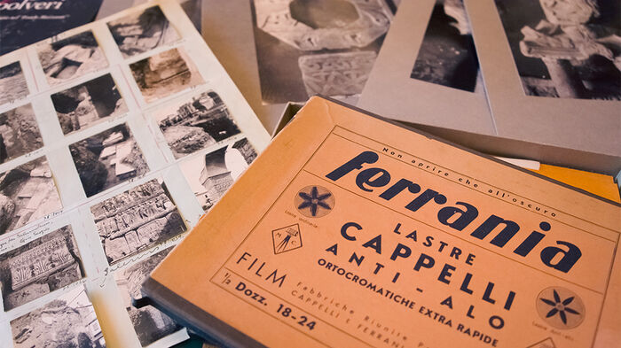 On a table is a brown, older box for photographs that say in Italian: "Ferrania. Loading Cappeli". All around are black and white photographs.