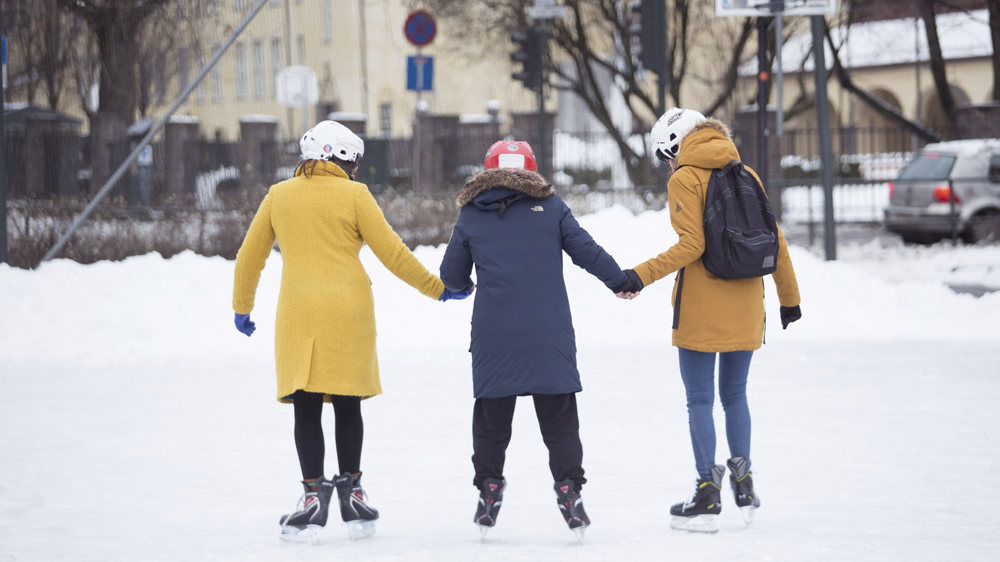 Three women holding hands on  an ice skating rink, wearing ice skates, helmets and winter clothes