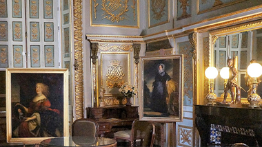Interior of a well-decorated room with portrait painting and gold details. The parquet is made of dark and light wood laid in a pattern, and it is high under the ceiling. Photo.