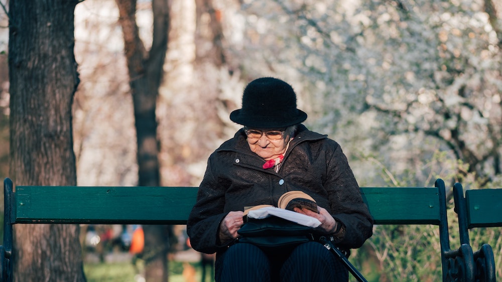 An old woman reading on a bench in a park. 