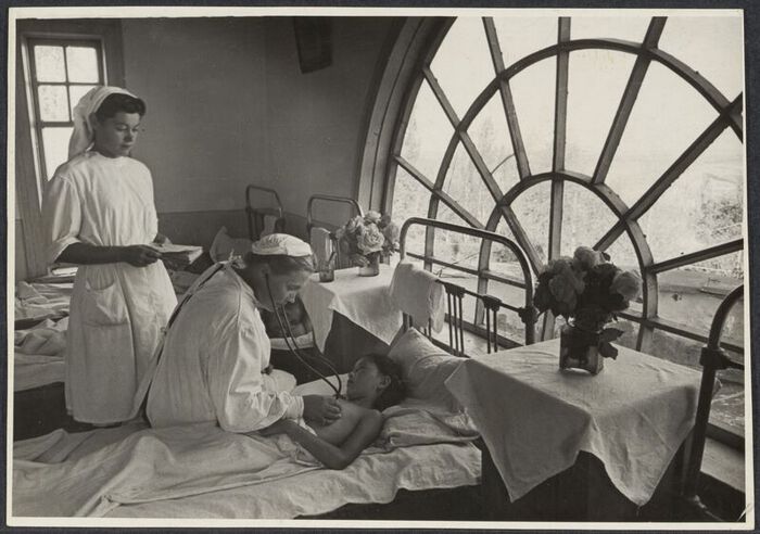 A black and white photograph showing two nurses examining a child lying in bed at the The Cholpon-Ata Children's Sanatorium in the Kirghiz Soviet Socialist Republic.