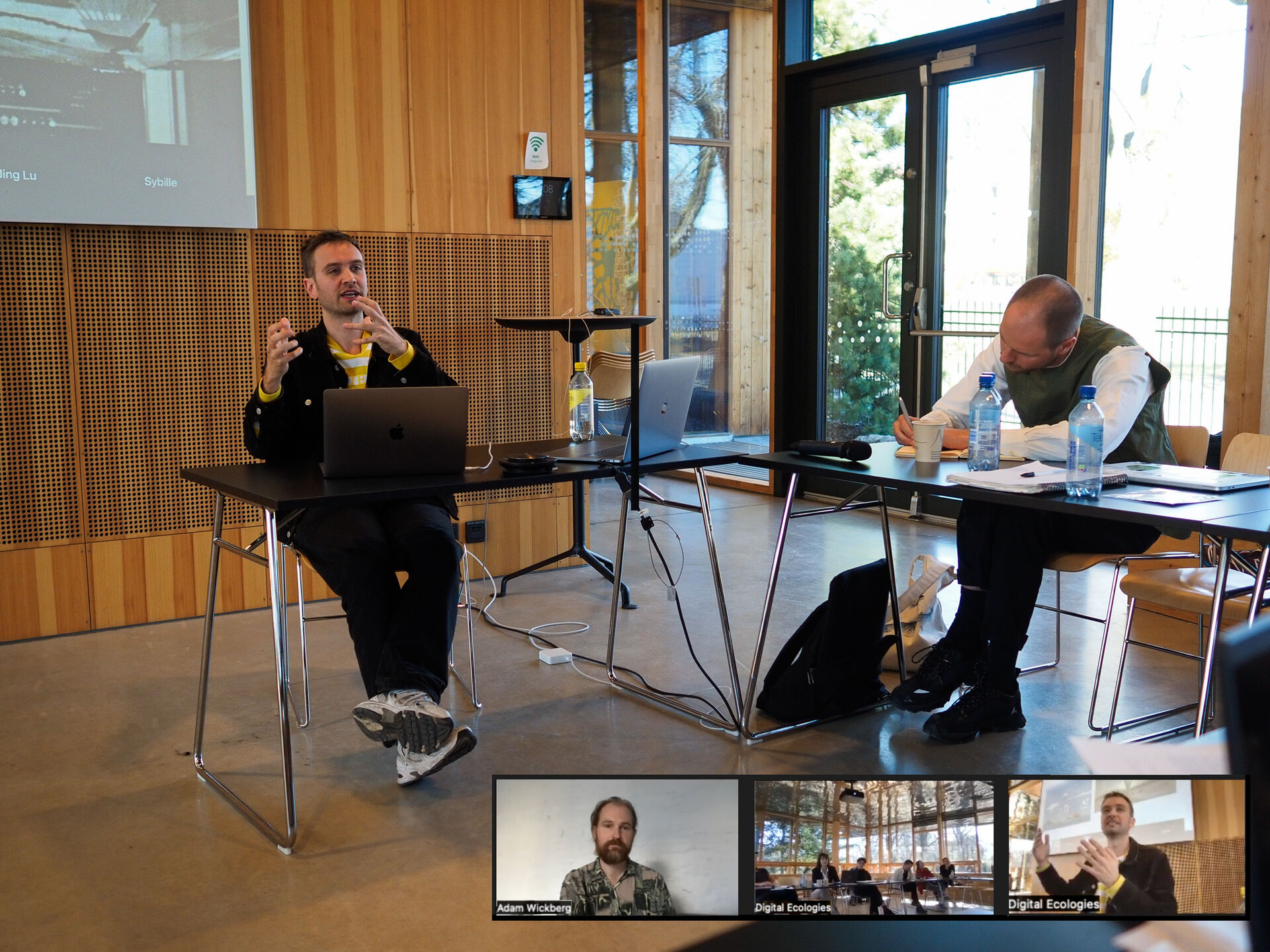 Screenshot of a Zoom meeting. A man is seated at the front of the room presenting. On his side is a member of the audience in the room, and you can also see small images of people attending virtually. 