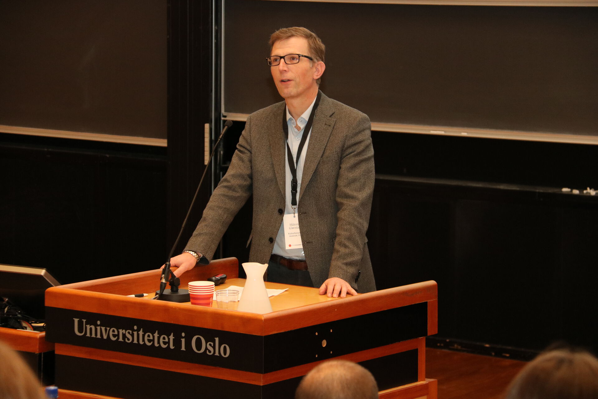 Mann i lys skjorte og mørk dressjakke bak en talerstol i en forelesningssal med påskriften "Universitetet i Oslo." 