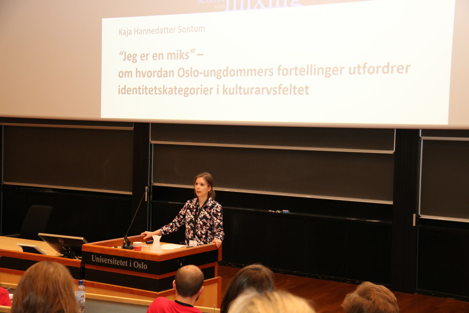 Kvinne i blomstrete skjorte bak en talerstol i en forelesningssal med påskriften "Universitetet i Oslo." I bakgrunnen en stor skjerm med teksten "Jeg er en miks" - om hvordan Oslo-ungdommers fortellinger utfordrer identitetskategorier i kulturarvsfeltet.