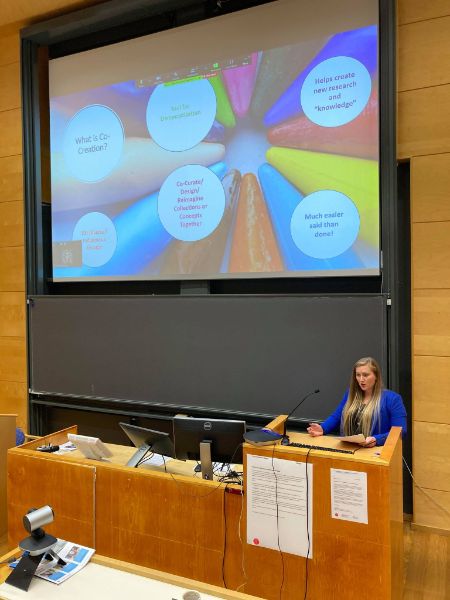 Person på en talerstol foran en stor skjerm i en forelesningssal. Foto.