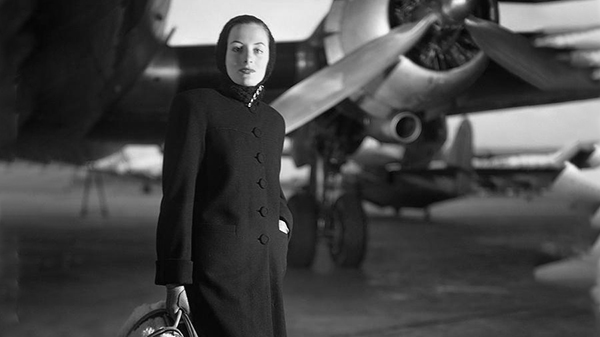 Black and White photo of stylish woman standing in front of an airplane.