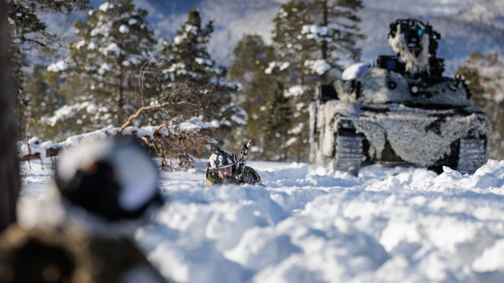 To soldater ligger i snøen og sikter mot hverandre. Bak den ene soldaten står en stridsvogn. Foto.