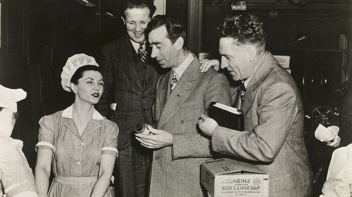 Three men in suits talking to a woman in a dress and hair net in a factory room. In the foreground a stack of cardboard boxes with "Heinz" written on them. Black and white photography.