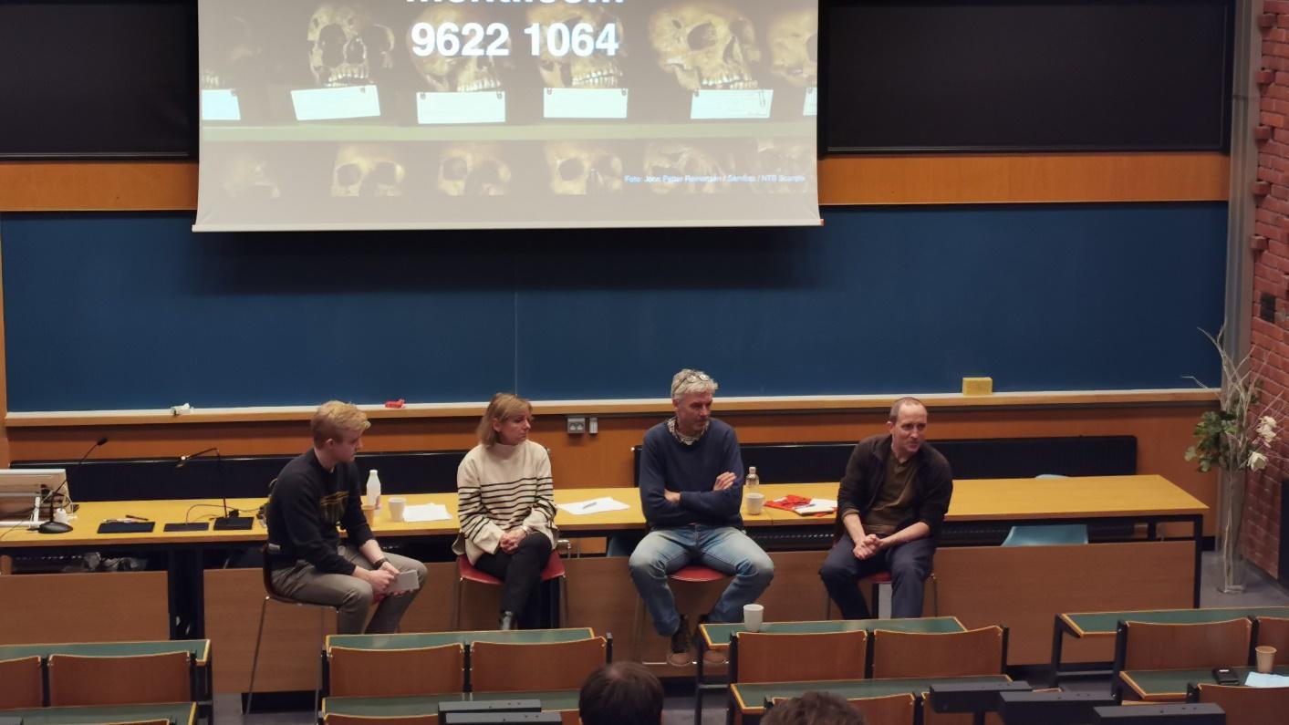 En gruppe mennesker sitter oppå pult i et auditorium. Bak gruppen vises en skjerm med bilder av ulike hodeskaller.