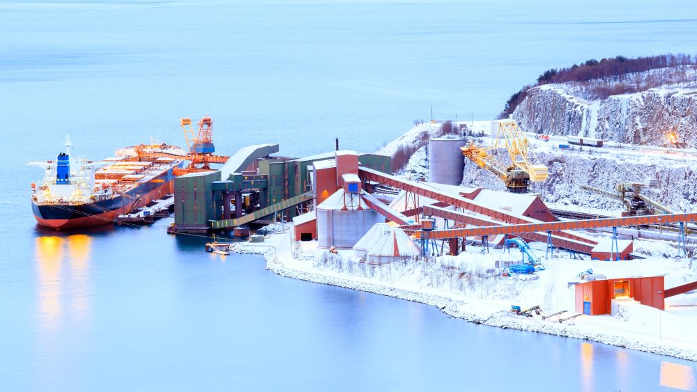 Stort containerlasteskip som ligger ved lastekai. På kaien er det flere store installasjoner for lasting av containere, og bakken rundt kaien er dekket av snø. Foto.