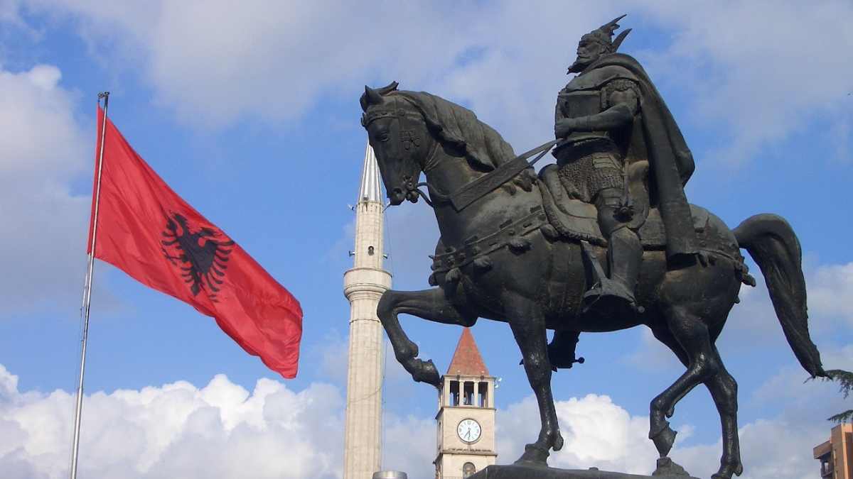 Foto av en mørk statue av en mann i rustning som sitter på en hest, men blå himmel, et tårn og et rødt flagg i bakgrunnen.