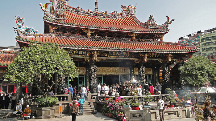 A Buddhist-Daoist temple with red-tile roof and dragon statuettes. Trees and people in the foreground.