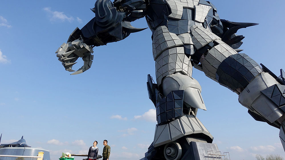 A giant robot stands beside two men with clear blue sky in the background.