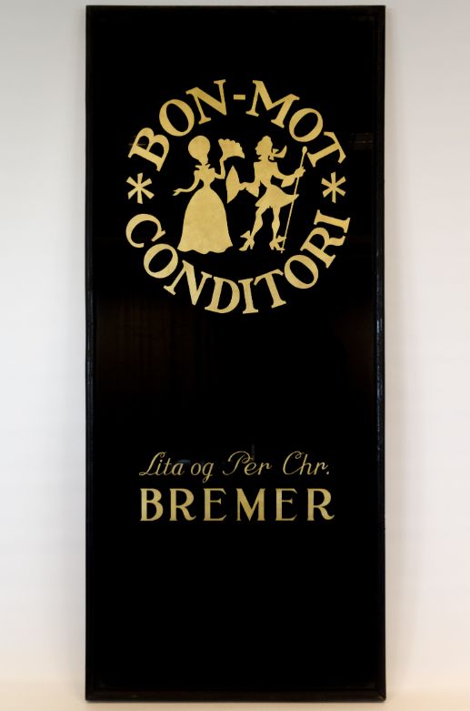 Svart skilt der det står med skrift i gull "Bon-Mot Conditori" og en stilisert kvinne og mann i pene klær. Lenger ned står det "Lita og Per Chr. Bremer".