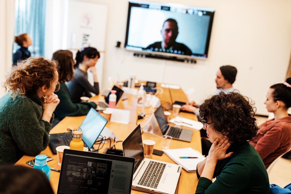 Students sitting around a table in front of their laptops, big screen showing other students on Zoom