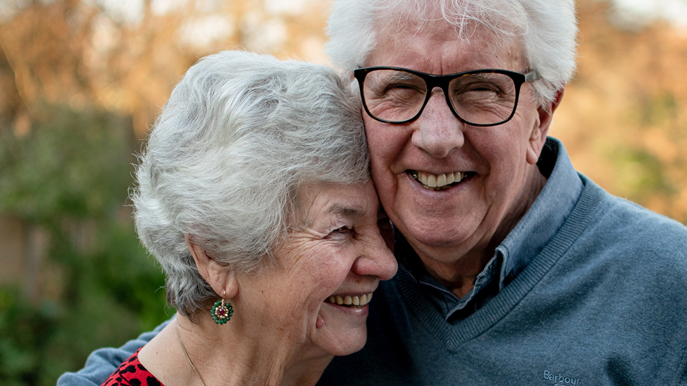 En eldre mann smiler mot kameraet mens han holder rundt en eldre dame. Foto. 