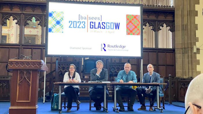 Four people sitting behind a table on a stage with a big screen in the baclgroun saying "2023 Glasgow"