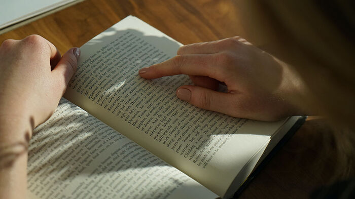 Bok med åpen side og en person som leser og holder fingeren sin på teksten