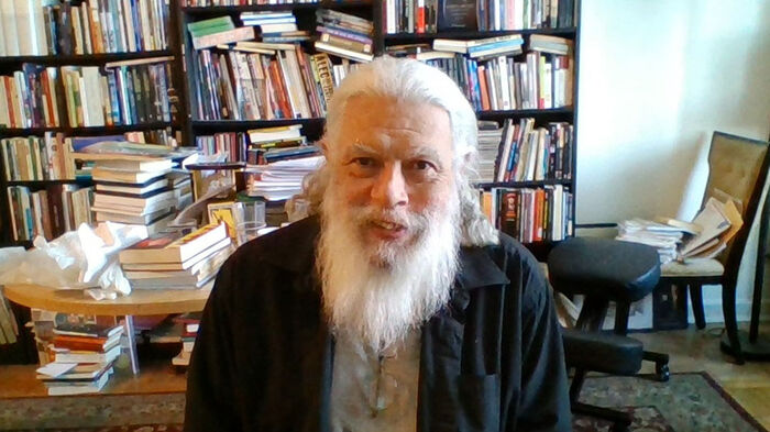 En mann med hvitt hår og skjegg som ser inn i kamera, i bakgrunnen en stor bokhylle. Foto.