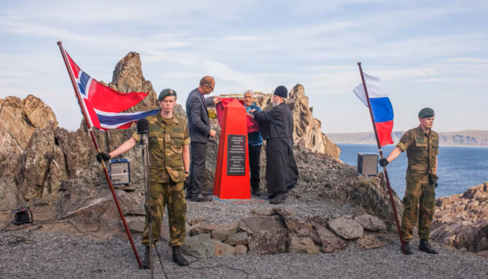 To norske soldater står på en steinrøys ved havet og holder i et norsk og et russisk flagg. Bak står tre menn og tar avdekket av en 1,5 meter høy, rød byste.