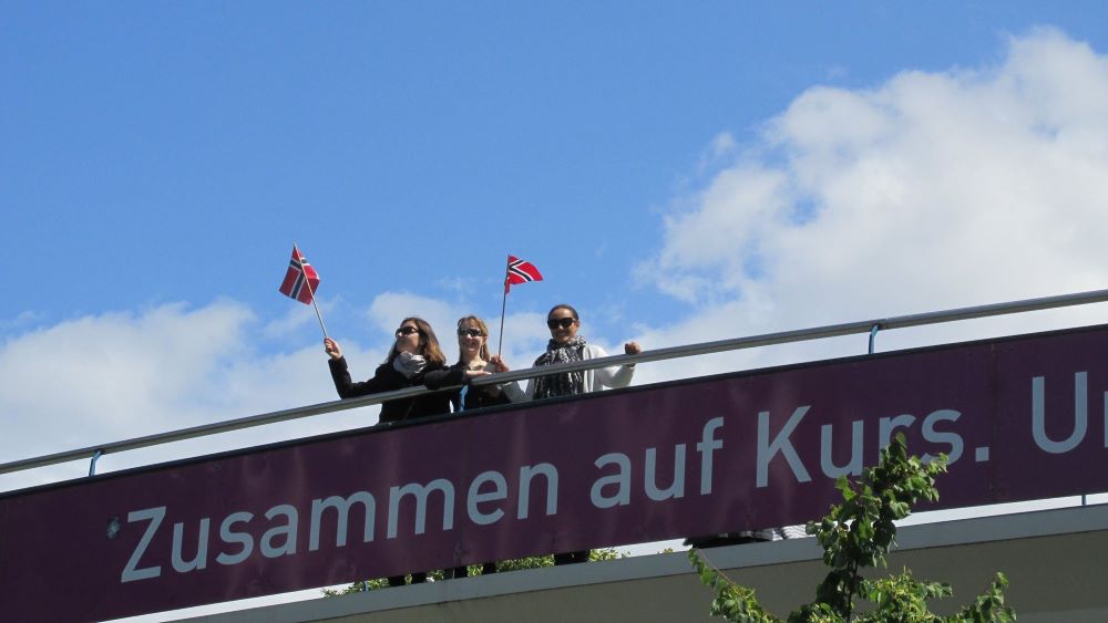 Tre personer på en bru som vinker med norske flagg. På brua er det en banner med påskriften "Zusammen auf Kurs"