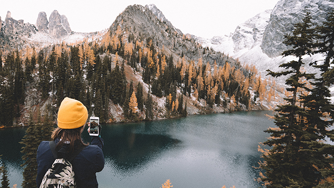 A beautiful vista of a lake, a forest and snowcovered maountains, a person in front taking a photo with her phone