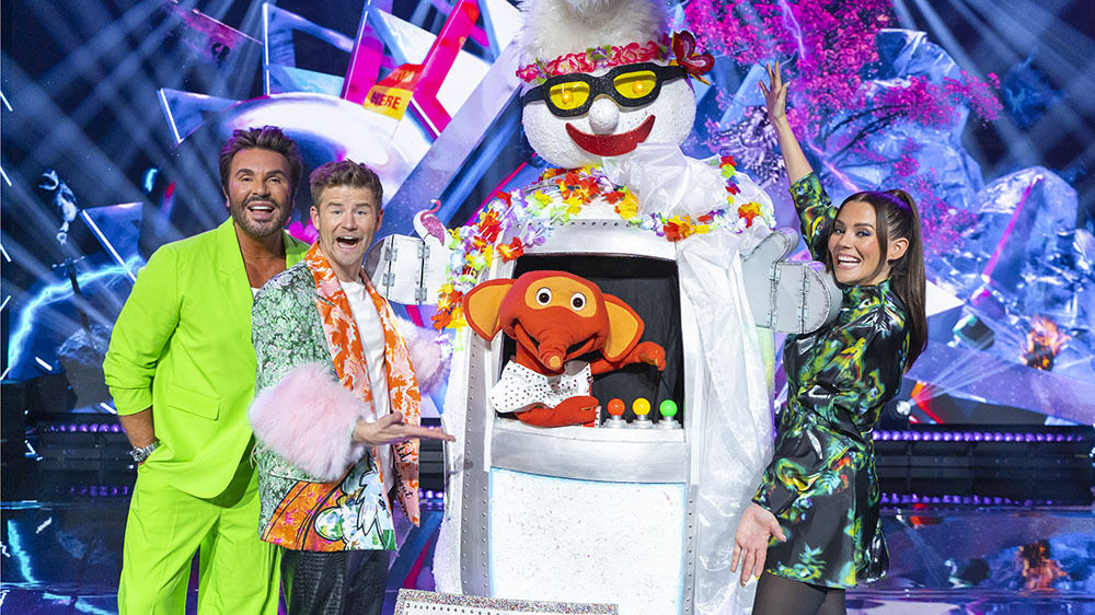 People in festive clothes are standing on a stage with a robot dressed as a snowman 