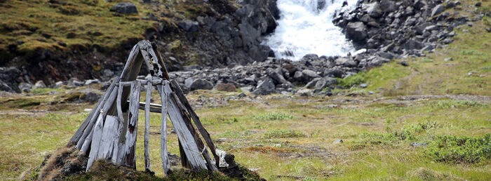 Rester etter en trehytte plassert i nordnorsk natur. I bakgrunnen renner en elv. Foto.