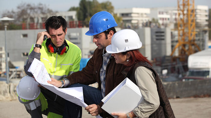 To mannlige bygningsarbeidere og en kvinne med hjelm ser på et kart eller dokument.