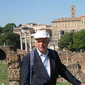 En eldre mann med dressjakke og hvit sixpence foran eldre bygninger i Roma. 