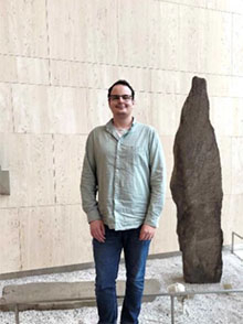 smilende mann med briller, blå jeans og lys grønn skjorte, stående i et museumslokale ved siden av en runestein