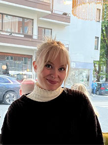 portrettfoto av smilende kvinne, mørk bakgrunn, blondt loppsatt hår, lys høyhalset under brun genser