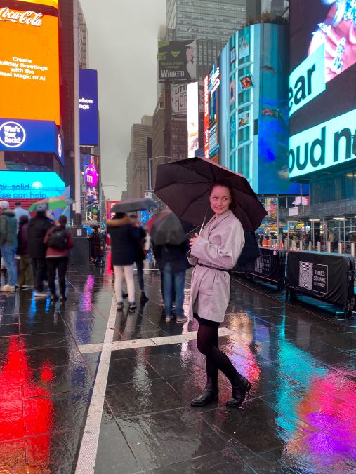 Gatebilde i regn, høye bygninger, kville i hvit kåpe under en paraply i forgrunn, mange mennesker i bakgrunn, neonfarger fra reklameskilt